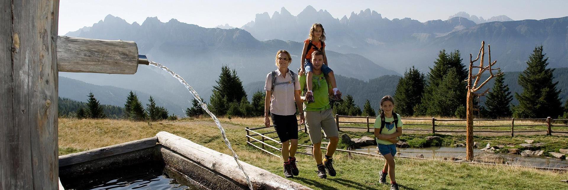 A family hiking past a fountain in the Dolomites, with the impressive peaks of the Odle group in the background.