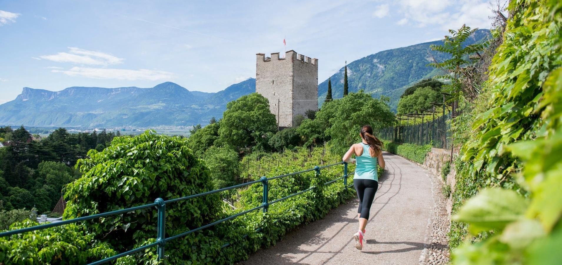 Eine Joggerin läuft auf einem Panoramaweg mit Blick auf einen Burgturm und Berge.