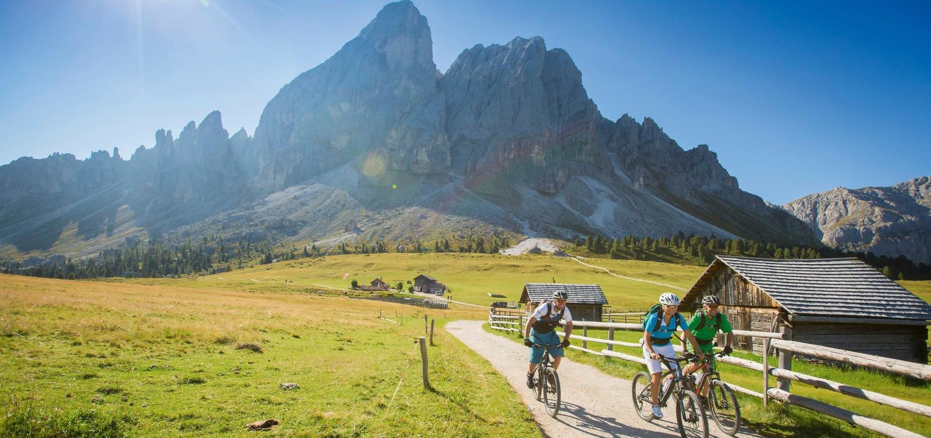 Drei Radfahrer auf einem Bergweg bei Sonnenschein mit Hütten und Dolomiten im Hintergrund.