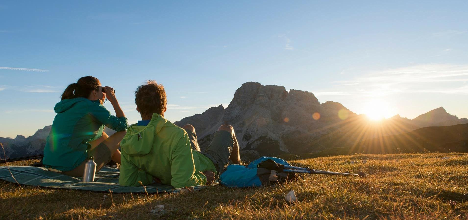 Zwei Wanderer sitzen auf einer Decke und schauen beim Sonnenuntergang auf die Berge.