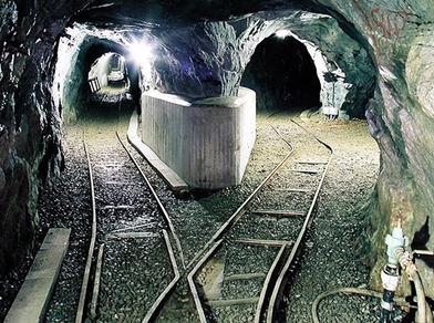Illuminated tunnel of a mine with tracks splitting in two directions. The walls are bare rock, and on the left side is a symbol with crossed hammer and pick.