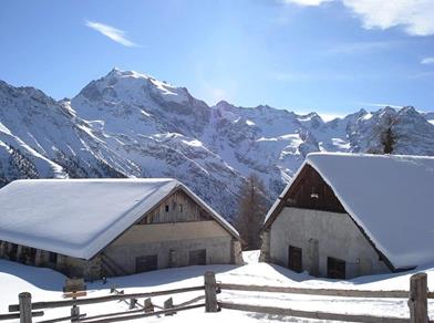 Zwei eingeschneite Berghütten vor beeindruckender Alpenkulisse bei strahlendem Sonnenschein.