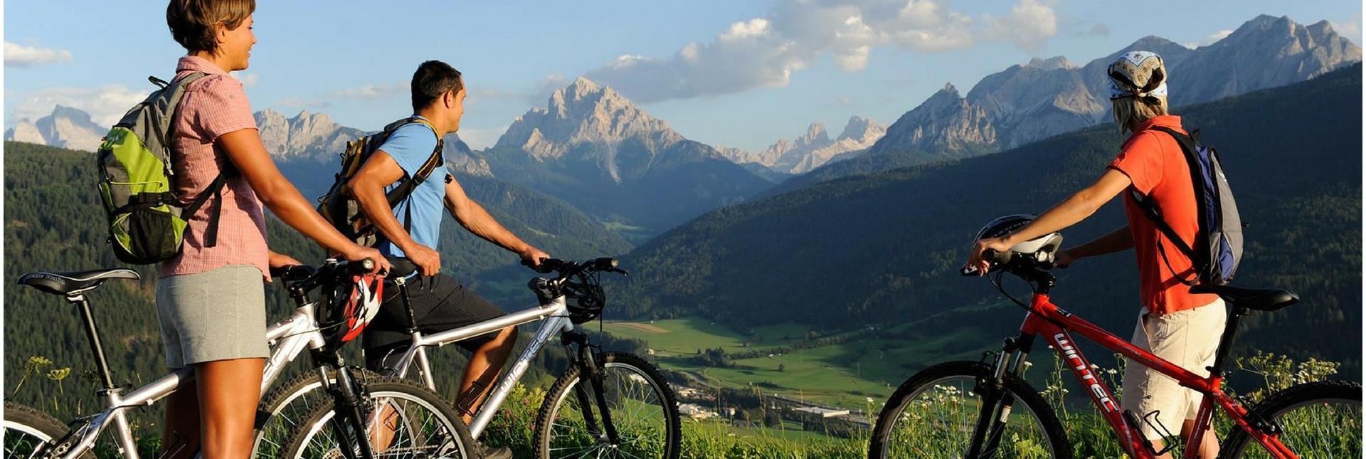 Drei Radfahrer mit Rucksäcken blicken von einem Aussichtspunkt auf ein grünes Tal und die Dolomiten.