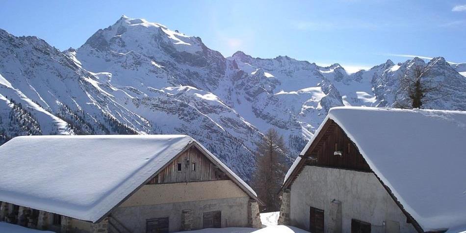 Zwei eingeschneite Berghütten vor beeindruckender Alpenkulisse bei strahlendem Sonnenschein.