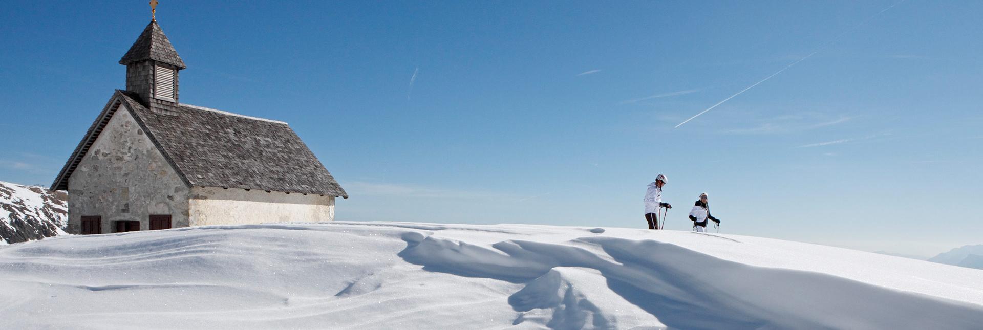Zwei Skitourengeher bei einer Kapelle in verschneiter Winterlandschaft unter blauem Himmel.